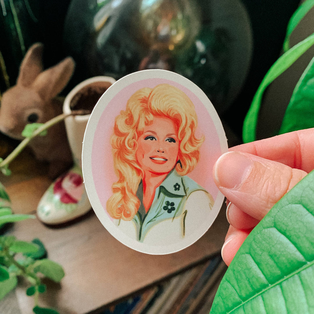 A hand holds an oval sticker featuring a retro-style illustration of a woman with blonde hair and a green shirt with a shamrock, set against a cozy, dark background with plants and a rabbit figurine.