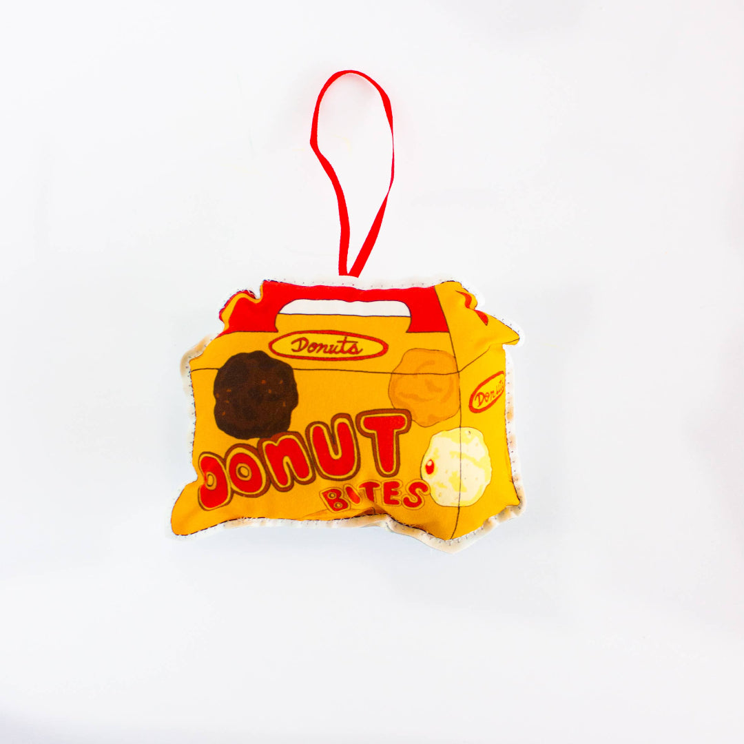 A close-up, high-angle flat lay shot on a plain white background features a soft, plush Christmas ornament shaped like a small, yellow and red box of "Donut Bites". The ornament has a red label that reads "Donuts" and a large red font that says "DONUT BITES". Three different colored donut holes are printed on the front of the box. A red ribbon loop is attached to the top for hanging.