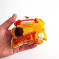 A close-up, high-angle flat lay shot on a plain white background features a soft, plush Christmas ornament shaped like a small, yellow and red box of "Donut Bites". The ornament has a red label that reads "Donuts" and a large red font that says "DONUT BITES". Three different colored donut holes are printed on the front of the box. A red ribbon loop is attached to the top for hanging.