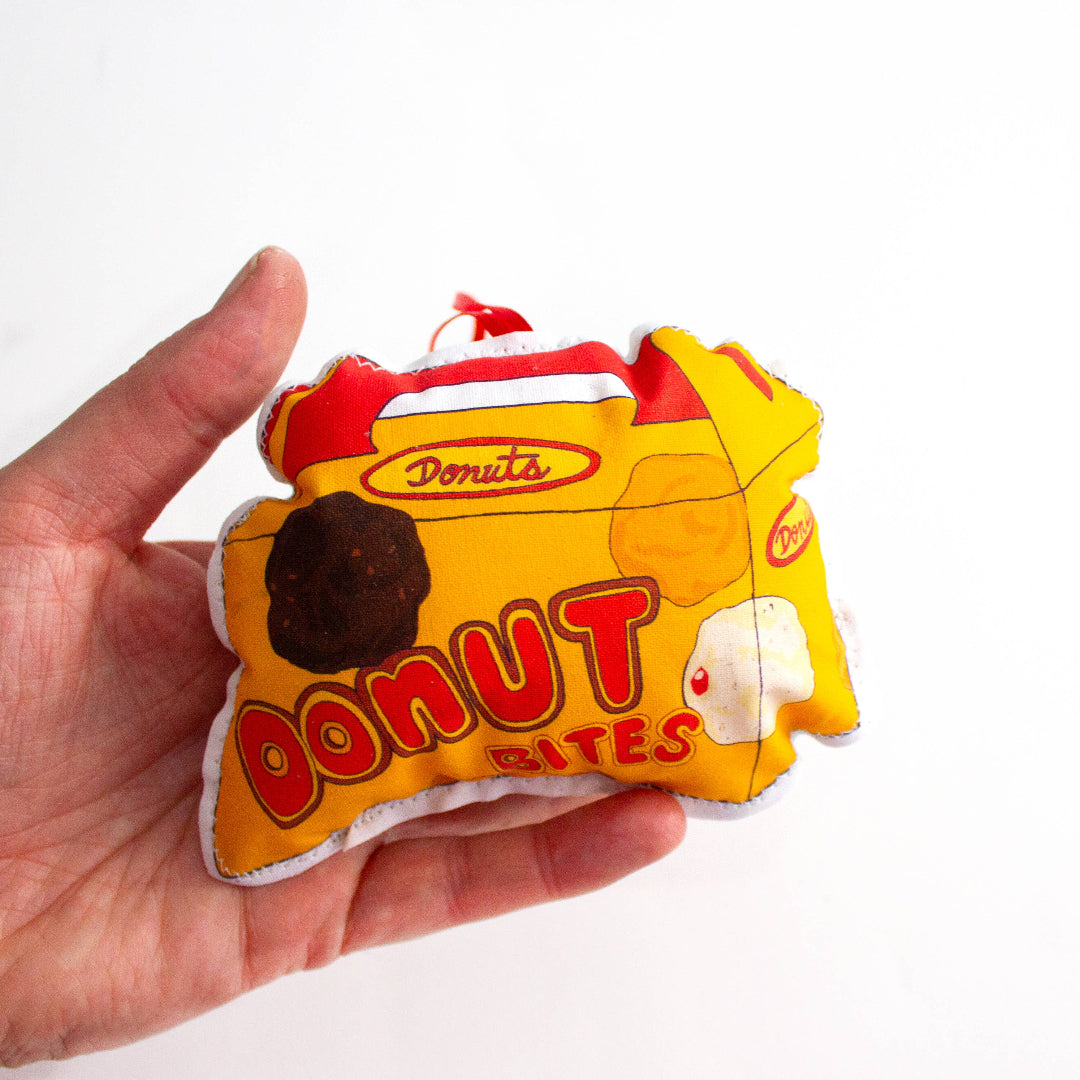 A close-up, high-angle flat lay shot on a plain white background features a soft, plush Christmas ornament shaped like a small, yellow and red box of "Donut Bites". The ornament has a red label that reads "Donuts" and a large red font that says "DONUT BITES". Three different colored donut holes are printed on the front of the box. A red ribbon loop is attached to the top for hanging.