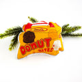 A close-up, high-angle flat lay shot on a plain white background features a soft, plush Christmas ornament shaped like a small, yellow and red box of "Donut Bites". The ornament has a red label that reads "Donuts" and a large red font that says "DONUT BITES". Three different colored donut holes are printed on the front of the box. A red ribbon loop is attached to the top for hanging.