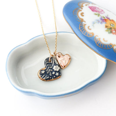A delicate 14K gold plated sterling silver 18-inch chain necklace featuring two small heart pendants made from imprinted porcelain. One heart is dark blue with white floral imprints and 22K gold detailing around the edge, while the other is a soft light pink with 22K gold detailing around the edge.