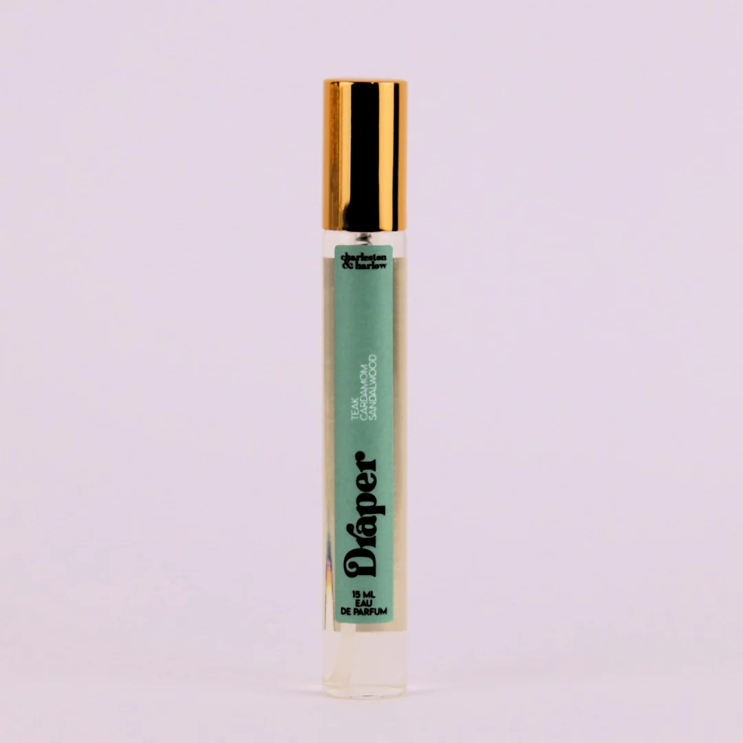 A small, cylindrical bottle of perfume with a gold cap. The bottle is transparent and filled with a light-colored liquid. The label on the bottle reads "Draper" in large, black letters. Below the brand name, it lists the fragrance notes: "Teak, Cardamom, Sandalwood."