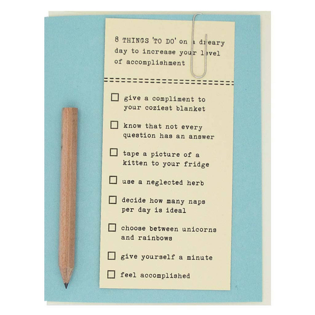 A light blue greeting card with a small, unsharpened pencil attached to the left side. On the right, a piece of cream-colored paper with a paperclip at the top lists "8 THINGS 'TO DO' on a dreary day to increase your level of accomplishment." The list includes: "give a compliment to your coziest blanket," "know that not every question has an answer," "tape a picture of a kitten to your fridge," "use a neglected herb," "decide how many naps per day is ideal," "choose between unicorns and rainbows," "give you