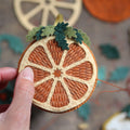 A close-up of hands holding a handcrafted felt ornament shaped like a dried orange slice, with green felt leaves on top, against a softly blurred rustic background.