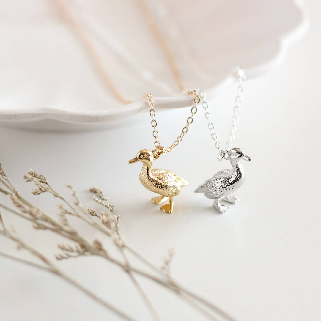 Two gold and silver duck-shaped pendants on necklaces with a soft background.
