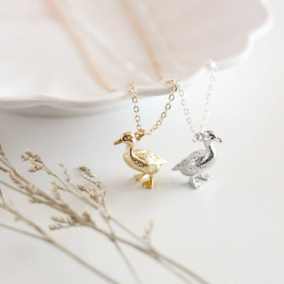 Two gold and silver duck-shaped pendants on necklaces with a soft background.