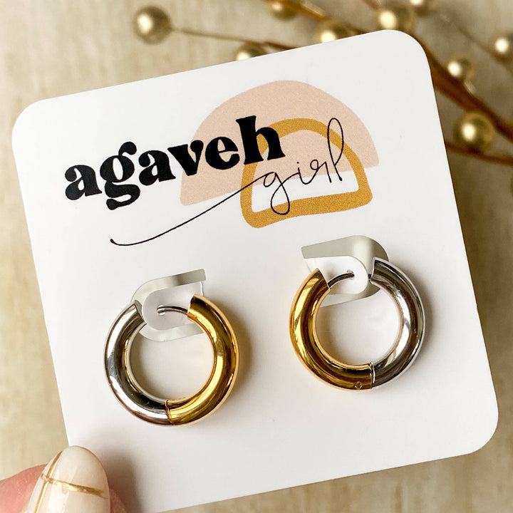 Two-tone chunky hoop huggie earrings (gold and silver) by agaveh girl, displayed on a branded card with the text 'agaveh girl,' against a textured background. The earrings are 4mm thick, made of stainless steel, and are sold in Ottawa, Ontario, Canada