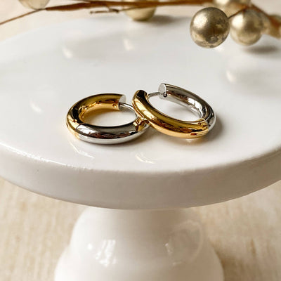 Two-tone chunky hoop huggie earrings (gold and silver) by agaveh girl, displayed on a branded card with the text 'agaveh girl,' against a textured background. The earrings are 4mm thick, made of stainless steel, and are sold in Ottawa, Ontario, Canada