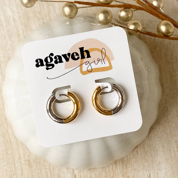 Two-tone chunky hoop huggie earrings (gold and silver) by agaveh girl, displayed on a branded card with the text 'agaveh girl,' against a textured background. The earrings are 4mm thick, made of stainless steel, and are sold in Ottawa, Ontario, Canada
