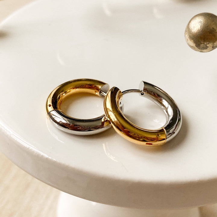 Two-tone chunky hoop huggie earrings (gold and silver) by agaveh girl, displayed on a branded card with the text 'agaveh girl,' against a textured background. The earrings are 4mm thick, made of stainless steel, and are sold in Ottawa, Ontario, Canada