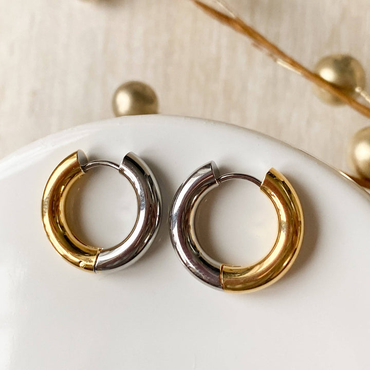 Two-tone chunky hoop huggie earrings (gold and silver) by agaveh girl, displayed on a branded card with the text 'agaveh girl,' against a textured background. The earrings are 4mm thick, made of stainless steel, and are sold in Ottawa, Ontario, Canada