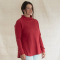 A woman wearing a red mock neck long-sleeve top and light blue jeans, standing against a neutral beige fabric backdrop.