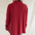 A woman viewed from behind wearing a red mock neck long-sleeve top and light blue jeans, standing against a neutral beige fabric backdrop.