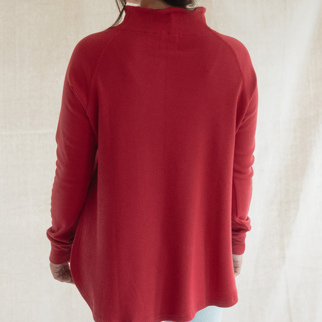 A woman viewed from behind wearing a red mock neck long-sleeve top and light blue jeans, standing against a neutral beige fabric backdrop.
