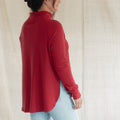 A woman wearing a red mock-neck long-sleeve top and light blue jeans, shown from a side-back angle against a neutral cream fabric backdrop.