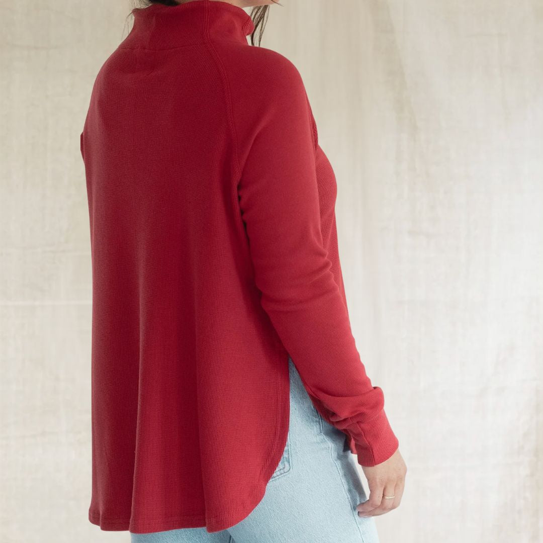 A woman wearing a red long-sleeve mock neck top and light blue jeans, shown from a side-back angle against a neutral textured beige background.