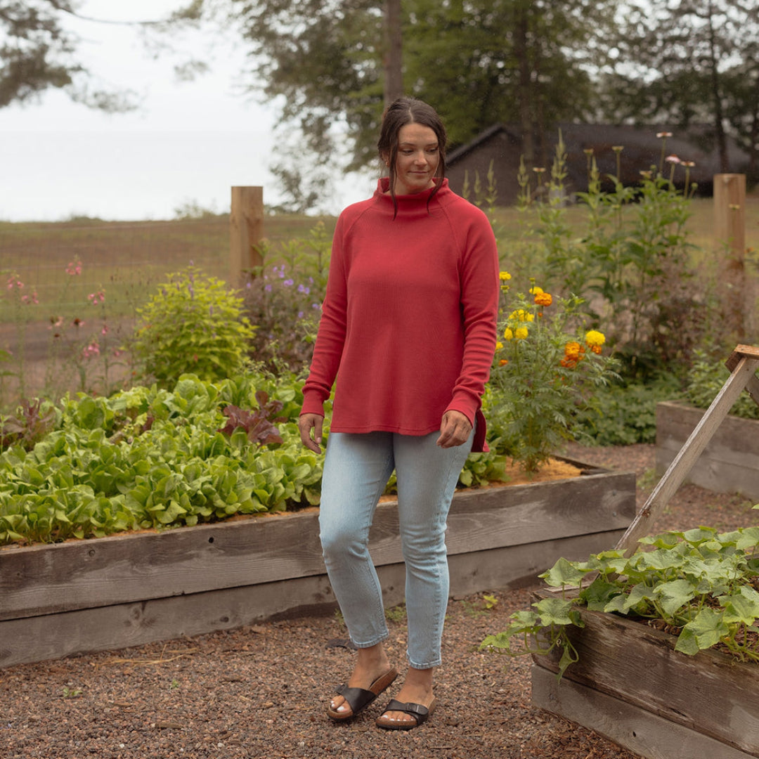 A woman wearing a red mock neck long-sleeve top, light blue skinny jeans, and black sandals stands in a gravel path of a rustic garden with wooden raised beds and green plants.