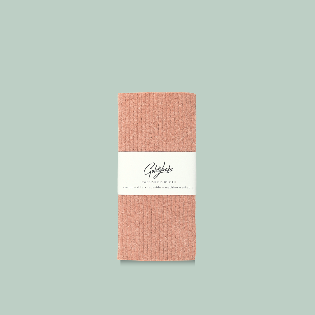A coral-colored Swedish dishcloth. The dishcloth has a textured surface and is labeled "Goldilocks Swedish Dishcloth" with information about its compostability, reusability, and machine washability.