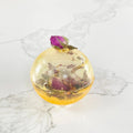A gold-colored tea bomb with a dried rose bud on top. The tea bomb is resting on a white marble surface.