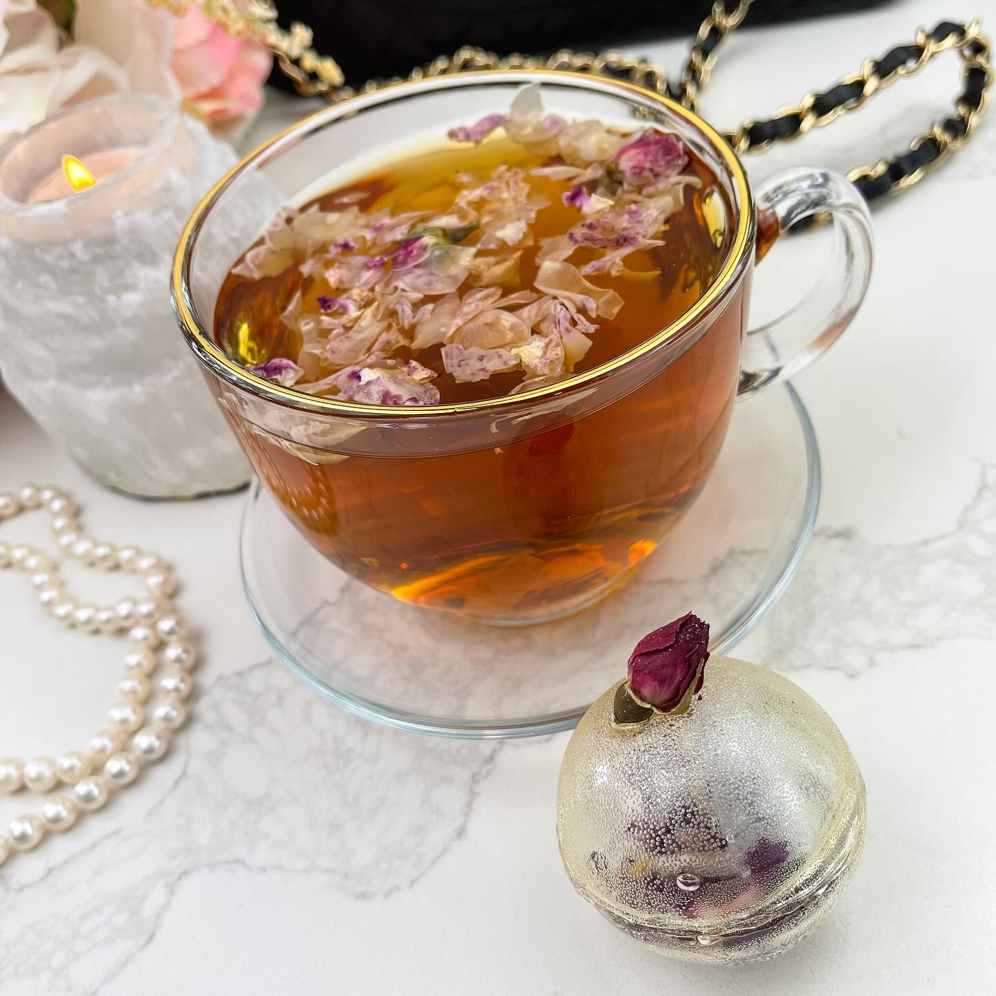 A gold-colored tea bomb with a dried rose bud on top. The tea bomb is resting on a white marble surface.