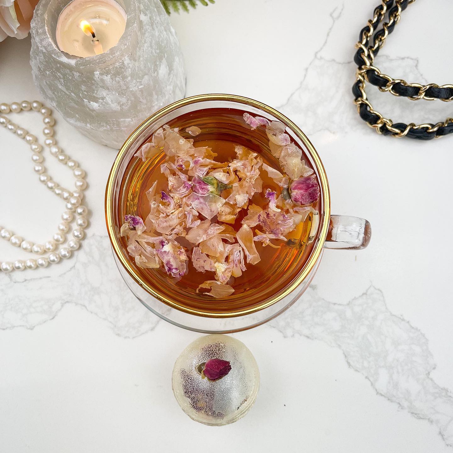 A gold-colored tea bomb with a dried rose bud on top. The tea bomb is resting on a white marble surface.