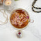 A gold-colored tea bomb with a dried rose bud on top. The tea bomb is resting on a white marble surface.