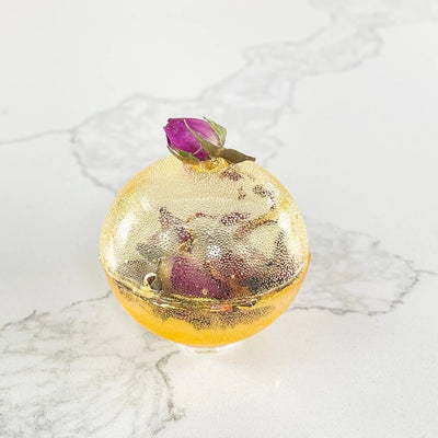 A gold-colored tea bomb with a dried rose bud on top. The tea bomb is resting on a white marble surface.