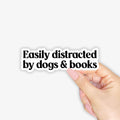 Hand holding a sticker with 'Easily distracted by dogs & books' text on a white background