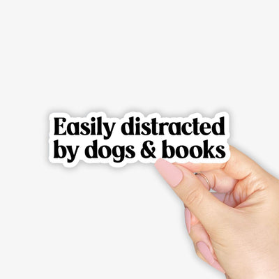 Hand holding a sticker with 'Easily distracted by dogs & books' text on a white background