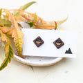 A pair of wooden earrings shaped like squares with half of each earring painted black and the other half a lighter color.