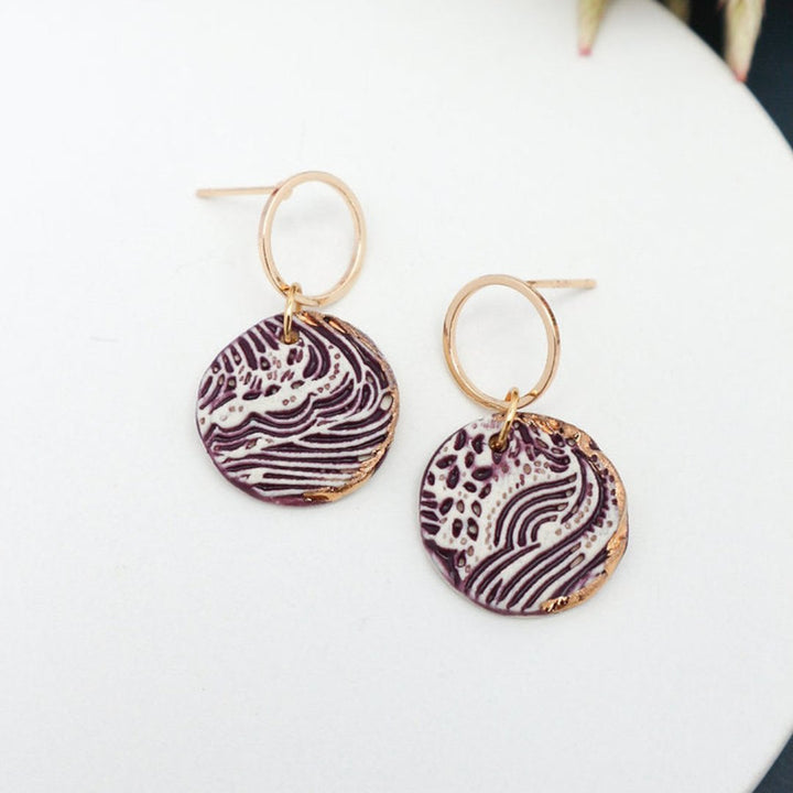 A pair of purple and white ceramic drop earrings with gold accents. The earrings feature a wave-like pattern and are suspended from gold hoops. They are displayed on a white circular plate with red flowers in the background.