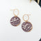 A pair of purple and white ceramic drop earrings with gold accents. The earrings feature a wave-like pattern and are suspended from gold hoops. They are displayed on a white circular plate with red flowers in the background.
