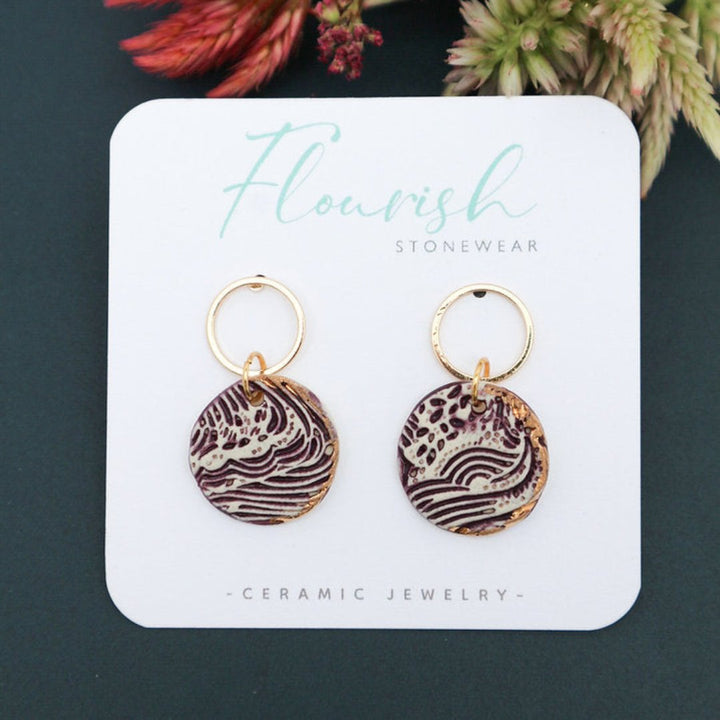 A pair of purple and white ceramic drop earrings with gold accents. The earrings feature a wave-like pattern and are suspended from gold hoops. They are displayed on a white circular plate with red flowers in the background.