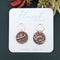 A pair of purple and white ceramic drop earrings with gold accents. The earrings feature a wave-like pattern and are suspended from gold hoops. They are displayed on a white circular plate with red flowers in the background.