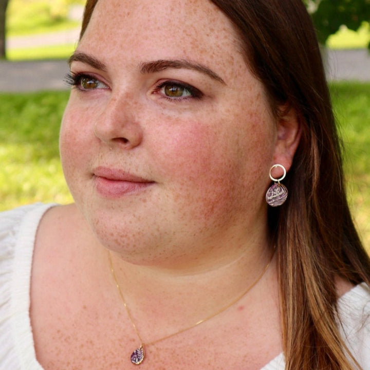 A pair of purple and white ceramic drop earrings with gold accents. The earrings feature a wave-like pattern and are suspended from gold hoops. They are displayed on a white circular plate with red flowers in the background.