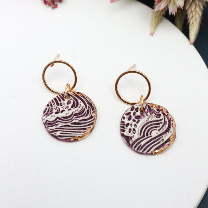 A pair of purple and white ceramic drop earrings with gold accents. The earrings feature a wave-like pattern and are suspended from gold hoops. They are displayed on a white circular plate with red flowers in the background.