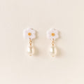 A pair of white flower-shaped dangle earrings with gold-tone metal accents and hanging pearl drops, displayed on a light cream background.