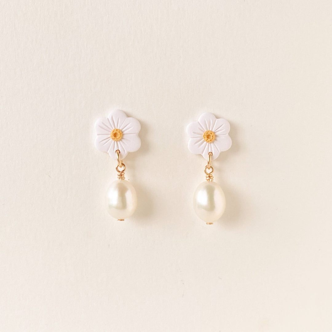 A pair of white flower-shaped dangle earrings with gold-tone metal accents and hanging pearl drops, displayed on a light cream background.