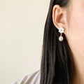 A close-up of a person's ear wearing a white flower-shaped dangle earring with a small gold accent and a dangling white pearl, against a plain light beige background.
