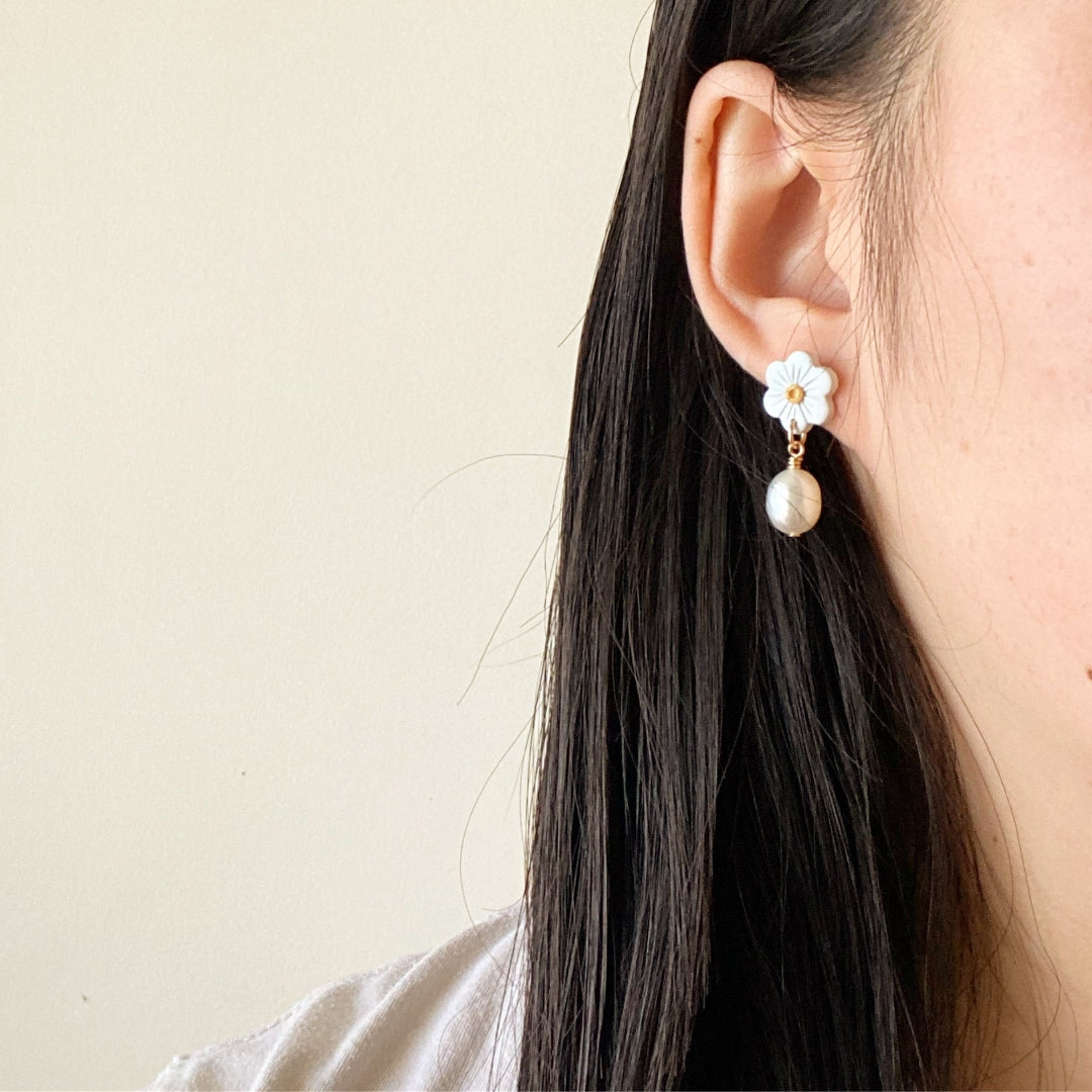 A close-up of a person's ear wearing a white flower-shaped dangle earring with a small gold accent and a dangling white pearl, against a plain light beige background.