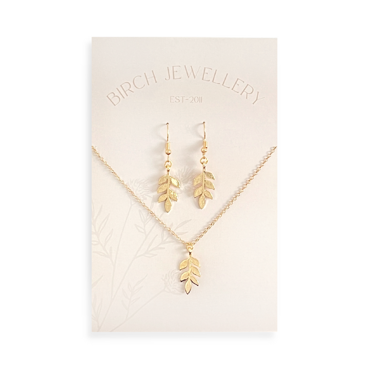 A gold necklace and earring set on a white card. The necklace features a gold leaf pendant, and the earrings are gold leaf-shaped drop earrings.