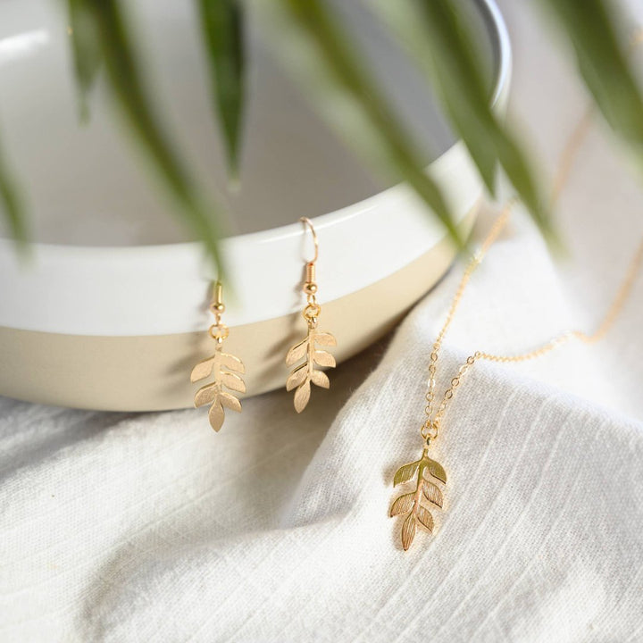 A gold necklace and earring set on a white card. The necklace features a gold leaf pendant, and the earrings are gold leaf-shaped drop earrings.