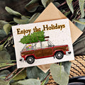 A card with the illustration of a old hatchback car with wood paneling with presents in the back and a tree strapped to the roof and the words "ENjoy the holidays"