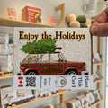A card with the illustration of a old hatchback car with wood paneling with presents in the back and a tree strapped to the roof and the words "ENjoy the holidays"