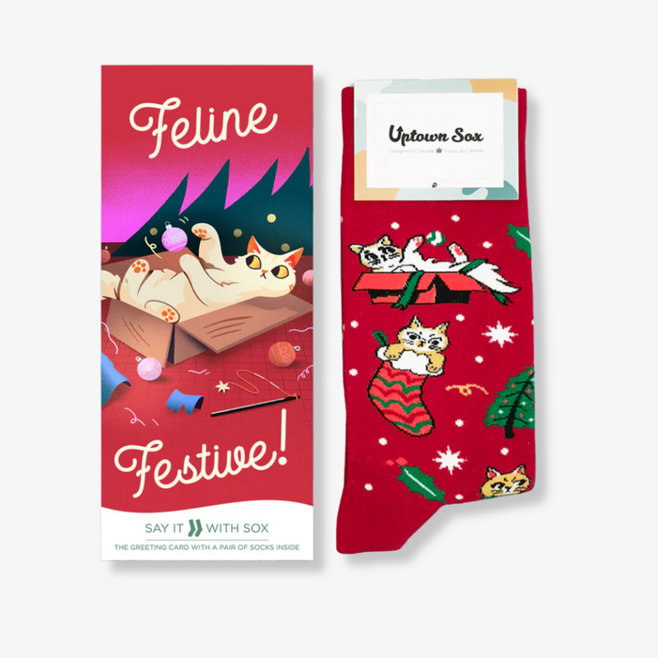 A pair of red socks with a festive cat design, packaged with a card. The socks feature cats playing with Christmas decorations, and the card has the text 