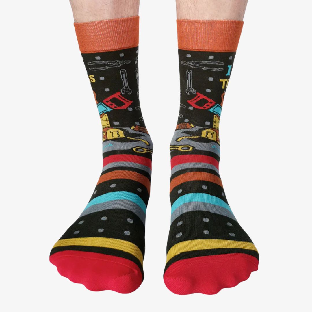 A pair of colourful men's crew socks featuring a black background with a pattern of small white dots, overlaid with horizontal stripes in red, blue, orange, yellow, and grey. One sock displays a cartoon illustration of a yellow toolbox overflowing with tools and the text "I FIX THINGS" in bright blue and yellow letters. The cuff of the sock is orange, the heel is yellow, and the toe is red. The brand "Uptown Sox" is subtly visible on the sole.