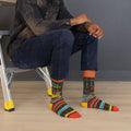 A pair of colourful men's crew socks featuring a black background with a pattern of small white dots, overlaid with horizontal stripes in red, blue, orange, yellow, and grey. One sock displays a cartoon illustration of a yellow toolbox overflowing with tools and the text "I FIX THINGS" in bright blue and yellow letters. The cuff of the sock is orange, the heel is yellow, and the toe is red. The brand "Uptown Sox" is subtly visible on the sole.