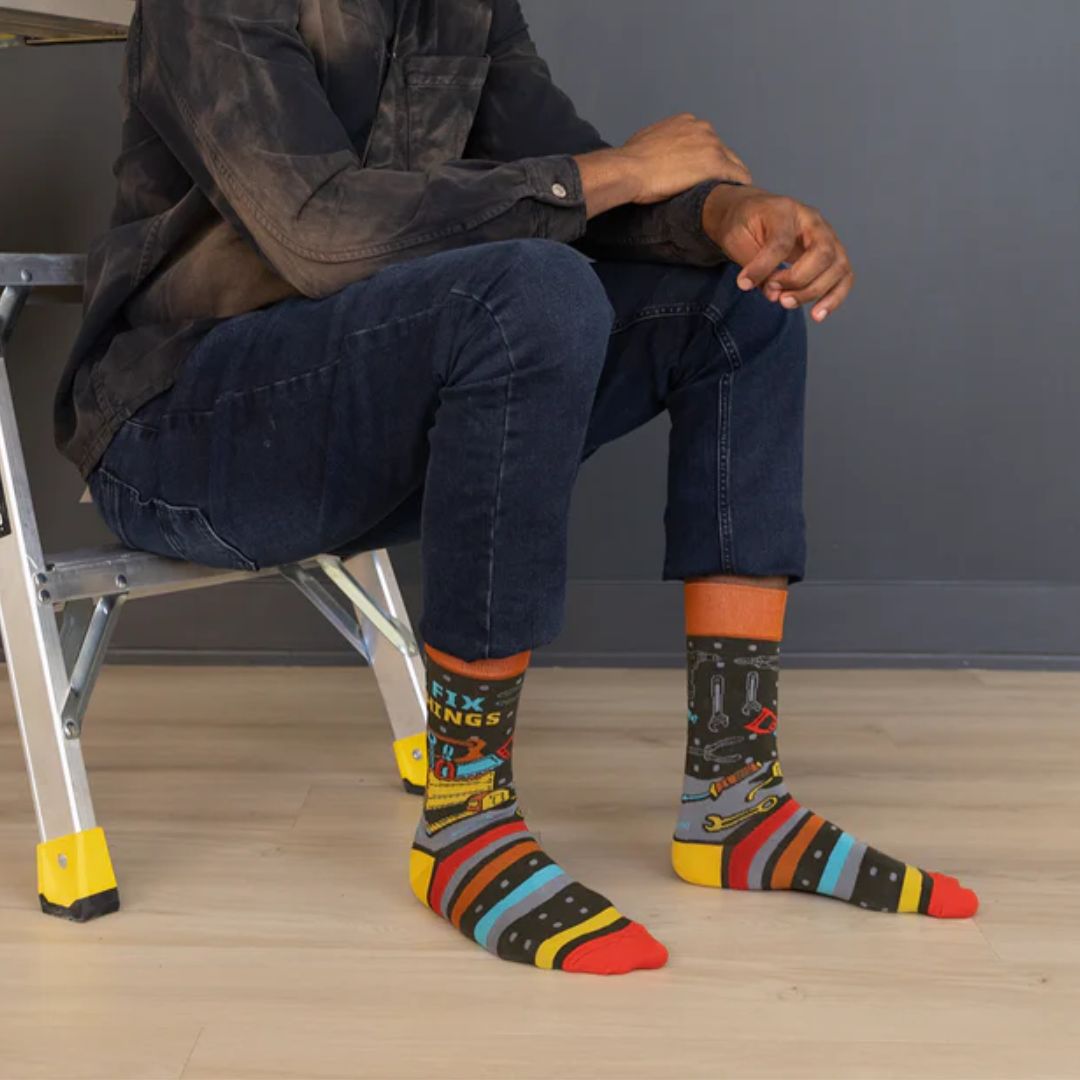 A pair of colourful men's crew socks featuring a black background with a pattern of small white dots, overlaid with horizontal stripes in red, blue, orange, yellow, and grey. One sock displays a cartoon illustration of a yellow toolbox overflowing with tools and the text "I FIX THINGS" in bright blue and yellow letters. The cuff of the sock is orange, the heel is yellow, and the toe is red. The brand "Uptown Sox" is subtly visible on the sole.