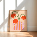 A framed art print featuring a retro-style illustration of orange flowers in a pink and orange striped vase, leaning against a white wall on a light wood floor with natural window shadows.